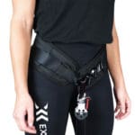 Wearing a black Exxentric Squat Hip Belt with metal carabiner and pulley system, a person in black leggings featuring a small logo on the thigh stands against a plain background.