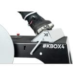 Close-up of a person using an Exxentric kBox4 Foot Block wearing black sneakers. The foot rests on the machines grey platform, featuring a black and white color scheme. #KBOX4 is visible on the side.