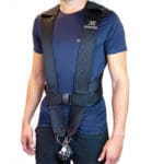 A person in a black Exxentric Squat Harness, featuring adjustable straps and a front buckle, with a metal attachment point at the bottom. They are wearing a navy blue T-shirt and dark pants.
