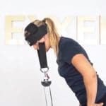 Someone wearing the Exxentric Head Harness is connected to a cable pulley system, performing a neck exercise. They lean forward, engaging in their fitness routine against a plain background with partial text visible.
