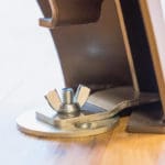 Close-up of the Exxentric kBox4 Floor Attachments metal wing nut and bolt on a flat plate mounted on wood, partially connected to a brown structure in the background, suggesting an adjustable mechanism.