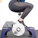 A person in black leggings and shoes squats on a kBox4 exercise device with EXXENTRIC and #KBOX4 markings. Their feet are on the platform, hands not visible. The scene includes the Exxentric Decline Board.