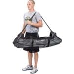 A person is standing, holding a large black mesh carrying bag with multiple handles, slung over one shoulder. The design is suitable for bulky items like sports equipment. The person is wearing a gray T-shirt, black shorts, and sneakers.