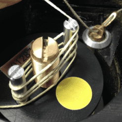 A close-up of the GymAware Tether: a mechanical device featuring a coiled wire around two metal posts, a central grooved brass piece, and a small yellow circle on a black background.