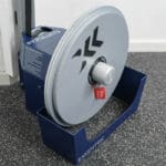 The Exxentric Advanced Flywheel Knob, a gray training device marked with kPulley2, features a large circular disc with black arrow designs. The floor beneath it is black speckled rubber.