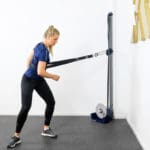 A woman in a gym uses the Exxentric Rotational Sling, performing resistance cable exercises. Shes wearing a blue shirt and black leggings, leaning slightly forward. The room features white walls and a carpeted floor.