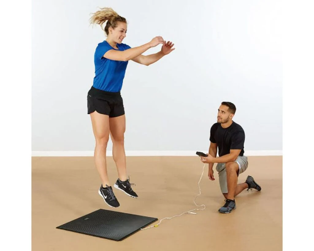 In minimal surroundings with a light brown floor, a woman in athletic wear jumps on the Just Jump Plyometric Upgrade mat as a man kneels beside her holding a connected device.