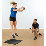 A woman jumps on a black mat while a man kneels nearby holding the Just Jump Plyometric Chip Upgrade device, monitoring her performance in a brightly lit indoor setting.