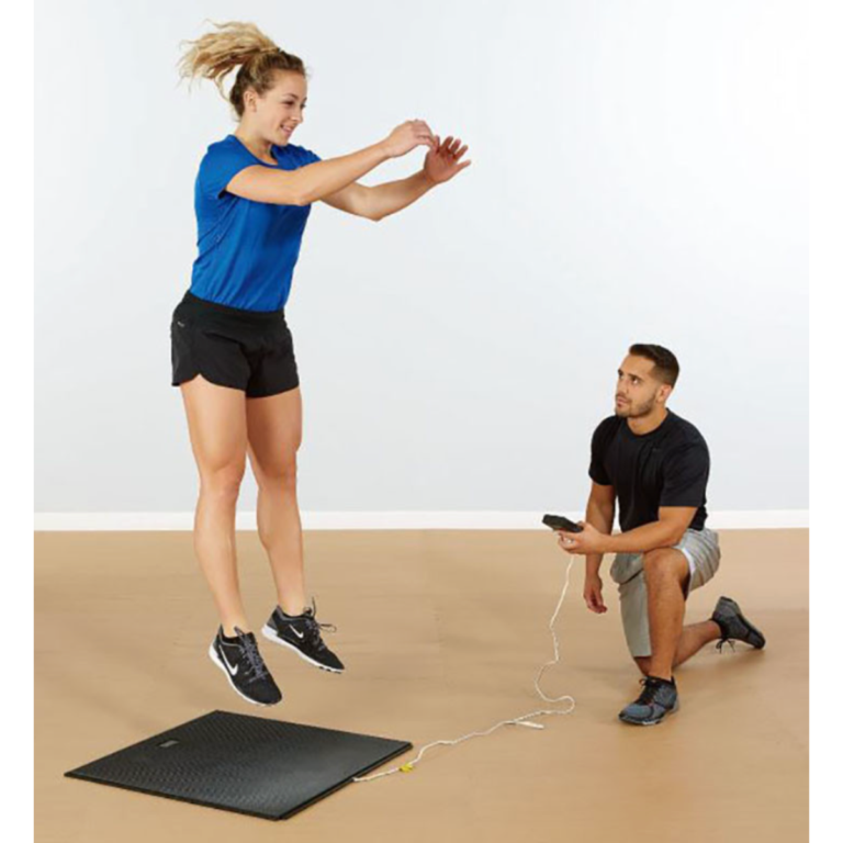 A woman jumps on a black mat while a man kneels nearby holding the Just Jump Plyometric Chip Upgrade device, monitoring her performance in a brightly lit indoor setting.