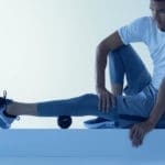 Wearing a white shirt and gray athletic pants, a person stretches their legs on a platform using the black Hyperice Hypersphere Mini massage ball under one thigh. The scene is highlighted with a cool blue tone, creating a serene and focused atmosphere.