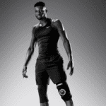 A man in athletic gear confidently poses against a gradient background, wearing a sleeveless top, sports shorts, and the Hyperice Ice ICT Compression Technology Knee brace on his right leg. The image has a sleek, dynamic black-and-white look.