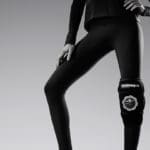 A person stands confidently with one hand on their hip, wearing a black outfit and a Hyperice Ice ICT Compression Technology Knee brace on their right leg. The gray backdrop highlights the sleek, modern look.