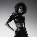 A woman with curly hair confidently poses for the camera in a sports bra and leggings, wearing a Hyperice Ice ICT Left Shoulder brace. The image is in black and white.