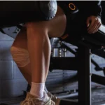 A person wears AirBands 2.0 on their knee while sitting on gym equipment, dressed in beige sneakers and white socks, with their hand resting on their thigh in a dimly lit fitness room.
