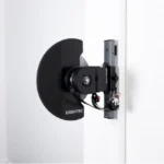 A black Exxentric kPulley GO, designed for resistance training, is mounted on a white wall. It has a compact design with a circular flywheel and electronic components attached.