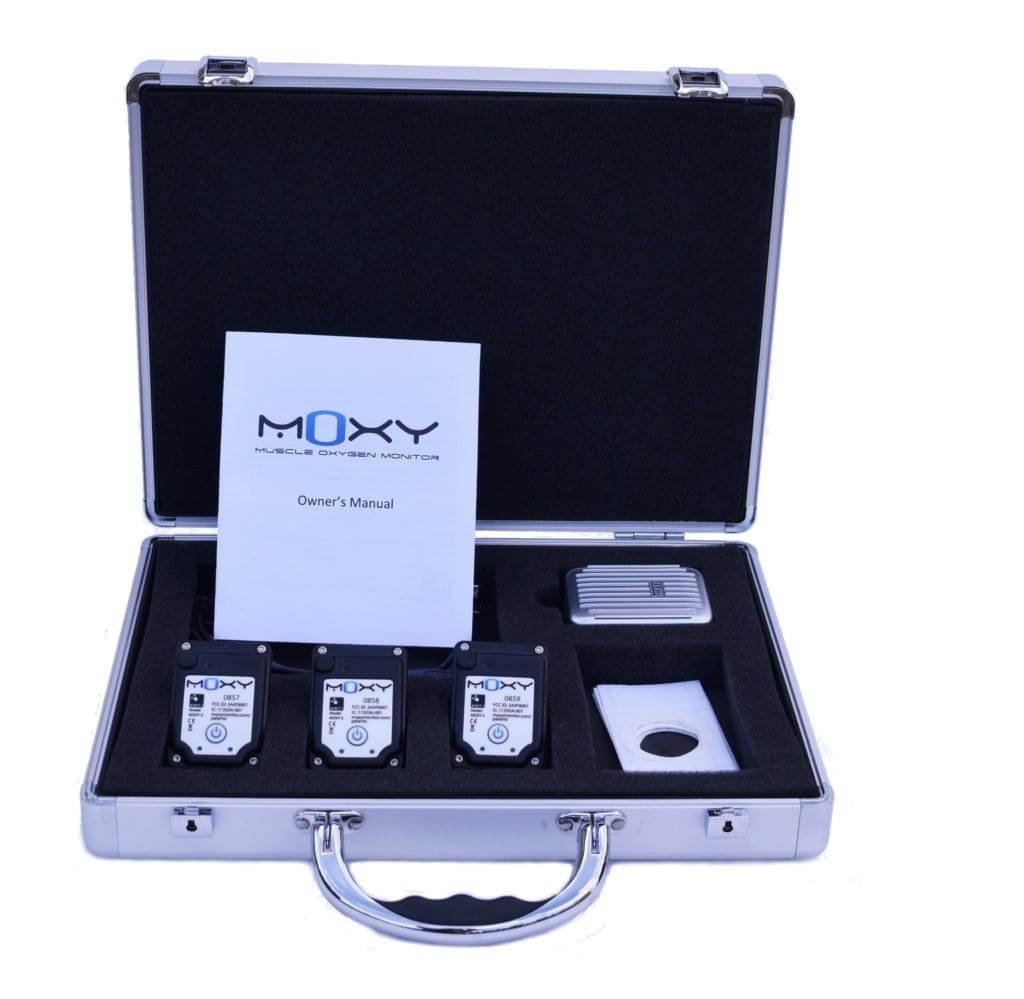 A silver briefcase is open, revealing a Moxy Muscle Oxygen Monitor set. Inside are three devices, a small rectangular unit, an owners manual, and other accessories, all fitted into a cushioned interior.
