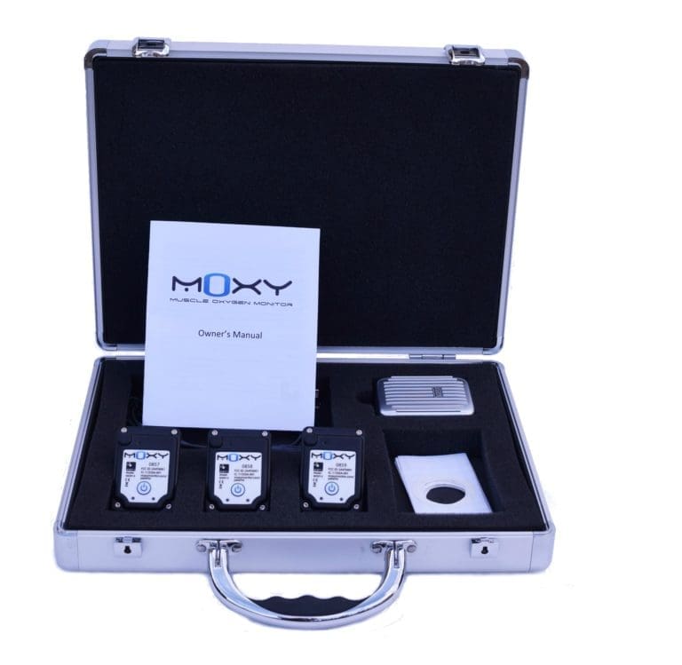 A silver briefcase is open, revealing a Moxy Muscle Oxygen Monitor set. Inside are three devices, a small rectangular unit, an owners manual, and other accessories, all fitted into a cushioned interior.
