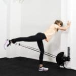 A woman in a peach shirt, black leggings, and white sneakers uses the Exxentric kPulley GO, extending one leg backward while holding a resistance band machine. She exercises on a black floor in a white room.