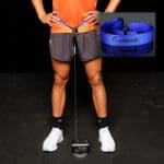 A person in an orange shirt and dark shorts stands on a black gym floor, legs shoulder-width apart. A blue resistance band connects from their waist to the floor. An inset shows the band close-up next to GymAware Jump Strap.