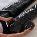 A person is lying on a bed wearing black compression leg sleeves from the Normatec 3 Full Body System. Theyre holding a smartphone in one hand and the devices control unit in the other, displaying Normatec branding.