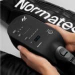 Someone holds a black controller for the Normatec 3 Lower Body System, showing digital settings with multiple buttons. Parts of the recovery boots can be seen in the background.