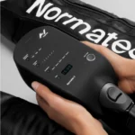 Someone holds a black controller for the Normatec 3 Lower Body System, showing digital settings with multiple buttons. Parts of the recovery boots can be seen in the background.