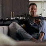 Someone sits on a couch wearing the Normatec 3 Full Body System sleeve on their arm, glancing at a smartphone in a modern room with a visible kitchen area in the background.