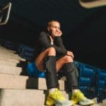 A woman in athletic gear sits on concrete steps in a dimly lit stadium, smiling. She sports the Normatec Go, complementing her black jacket, blue shorts, black knee-high socks, and yellow sneakers. Blue seats are seen behind her.