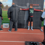 A person in athletic wear stands against a height measurement board with arms outstretched. Two individuals, one holding a clipboard, observe nearby on the running track where a Dashr Biostation is set up.