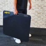 A person holds a large, black Skyhook Contact Mat with its yellow brand name visible. Theyre in casual attire on a dark carpeted floor, standing against a white brick wall.