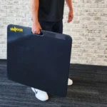 A person holds a large, black Skyhook Contact Mat with its yellow brand name visible. Theyre in casual attire on a dark carpeted floor, standing against a white brick wall.