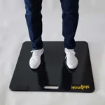 A person in white sneakers and dark jeans stands on a black Skyhook Contact Mat with Shhook in yellow at the corner, featuring a handle cutout.