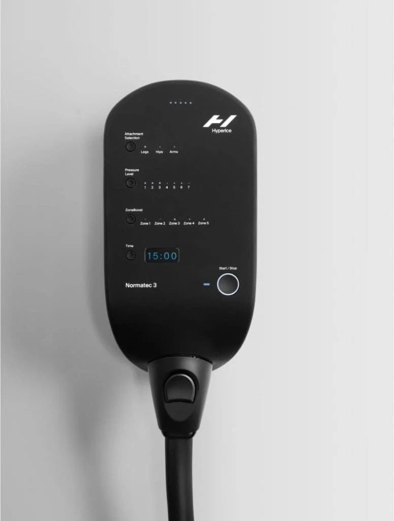 The image shows a black Normatec 3 Pump mounted on a wall. It has various buttons, LED indicators, and a display screen showing 15:00, with a cable extending downward from the devices bottom.
