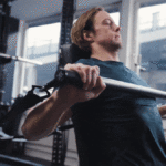 A person uses the Exxentric Upper Body Kit for seated cable rows in a gym, pulling a bar attached to the machine. Their upper back muscles engage as weightlifting equipment surrounds them, with natural light streaming through the windows.