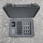 The Enode Pro – Charging Case lies open on concrete, featuring a textured protective lining. Inside are organized foam compartments with six small gadget slots, a rectangular item, and a coiled cable neatly stored.