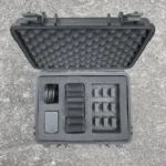 The Enode Pro – Charging Case lies open on concrete, featuring a textured protective lining. Inside are organized foam compartments with six small gadget slots, a rectangular item, and a coiled cable neatly stored.