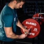 In a gym setting, a person wearing a teal shirt is preparing a barbell with red Eleiko weight plates, focusing on their hands and equipment like the Enode / Eleiko – Bar Sensor Kit.