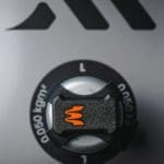 Close-up of the Enode Pro / Exxentric – Flywheel Adapter, featuring a black and silver machine knob with an orange emblem against a grey background, marked 0.050 kgm. Geometric shapes appear above.