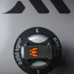 Close-up of the Enode Pro / Exxentric – Flywheel Adapter, featuring a black and silver machine knob with an orange emblem against a grey background, marked 0.050 kgm. Geometric shapes appear above.