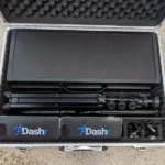A Dashr Display Pack rests in an open carrying case on a carpeted floor, showcasing its components: a large digital display, tripods, sensor devices, and accessories neatly arranged inside.