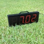 The Dashr Display is set on a grassy field, showing 7.02 in red. It features a black handle on top and has a visible power cord extending to the right.