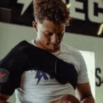 In a gym filled with equipment and motivational signs, a person with curly hair in a white shirt adjusts the Hyperice Venom 2 on their left shoulder while looking down.