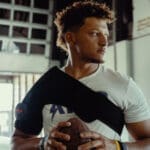 A person with curly hair is indoors holding a football and donning a Hyperice Venom 2 Right Shoulder brace. Theyre gazing to the side, clad in a casual white shirt, against a backdrop of large windows and industrial-style decor.