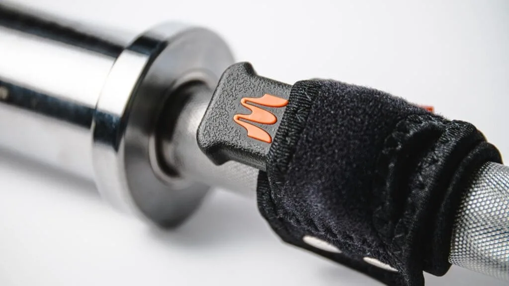 Close-up of a motorcycle handlebar grip with Enode Replacement Barbell Strap in black and orange attached to the throttle control. The grip has a textured surface, and the strap uses Velcro for secure fastening.