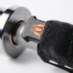 Close-up of a motorcycle handlebar grip with Enode Replacement Barbell Strap in black and orange attached to the throttle control. The grip has a textured surface, and the strap uses Velcro for secure fastening.
