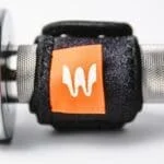 The Enode Replacement Barbell Strap, with an orange patch and white logo, secures the weight on a gym barbell with a textured metal grip against a plain, light background.