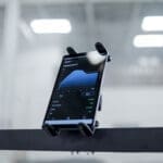 A SHREDmill smartphone, secured on a holder, shows a blue line graph on its dark screen. The background is an indoor area with blurred fluorescent ceiling lights.
