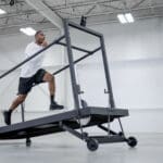 In an industrial-style gym, a person runs on the SHREDmill, a large inclined treadmill, beneath high ceilings and bright lighting. They sport a white shirt, black shorts, and black athletic shoes.
