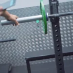 A person’s hand is placing or removing a barbell on the Vitruve Roller Hook squat rack, with a green weight plate and a patterned metal wall visible in the gym background.