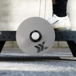 A person in black pants and white sneakers exercises on an Exxentric kBox Lite Starter System, featuring a central round pulley on a gray platform against a neutral background.